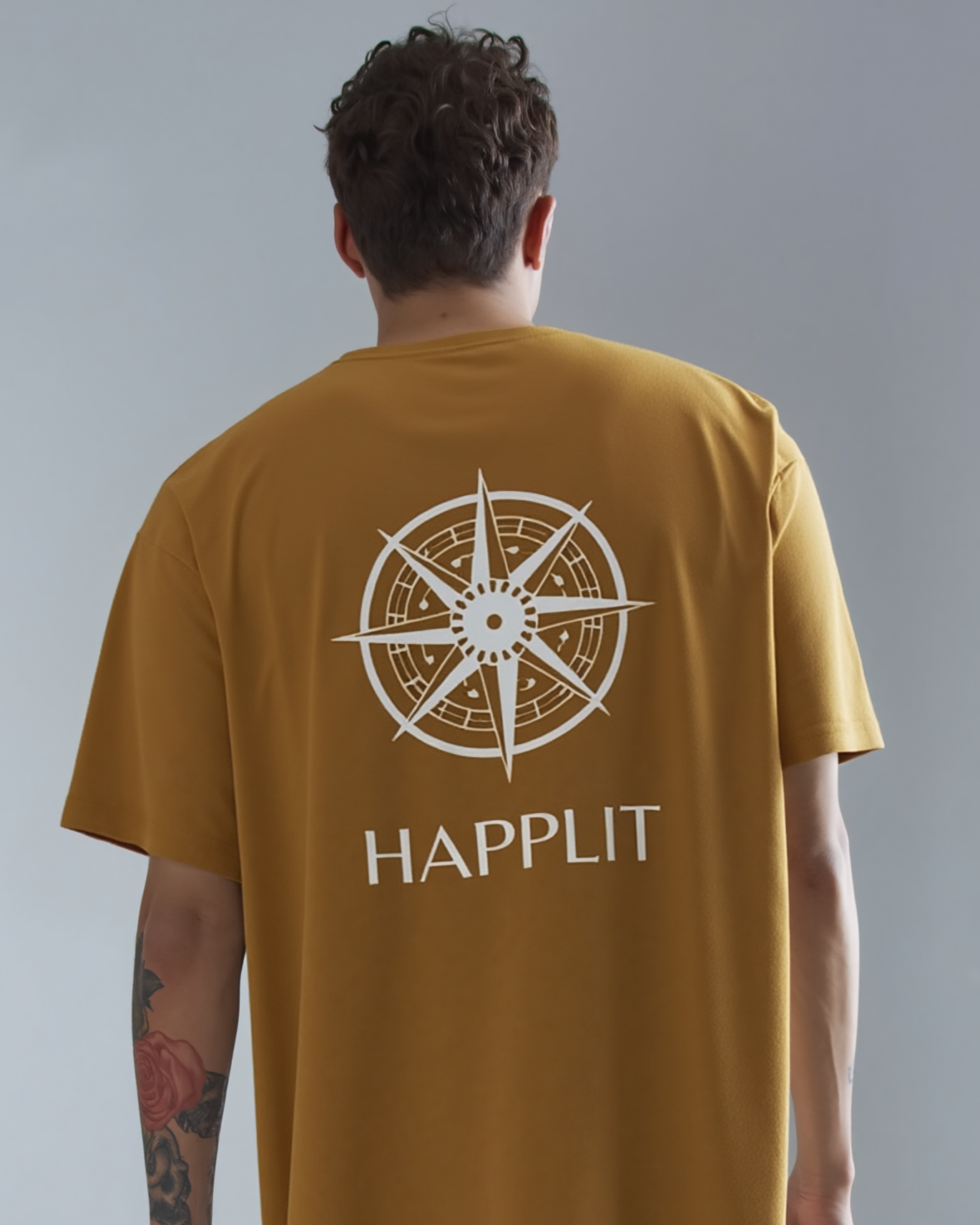 Person wearing a mustard yellow t-shirt with a compass design and 'HAPPLIT' text on the back, against a gray background.